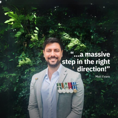 A man in a suit stands in front of foliage